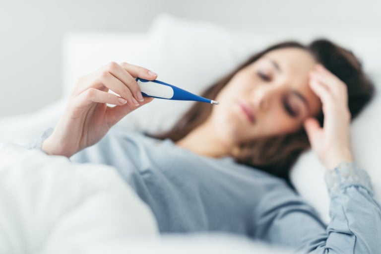 Woman in bed holding forehead and reading thermometer